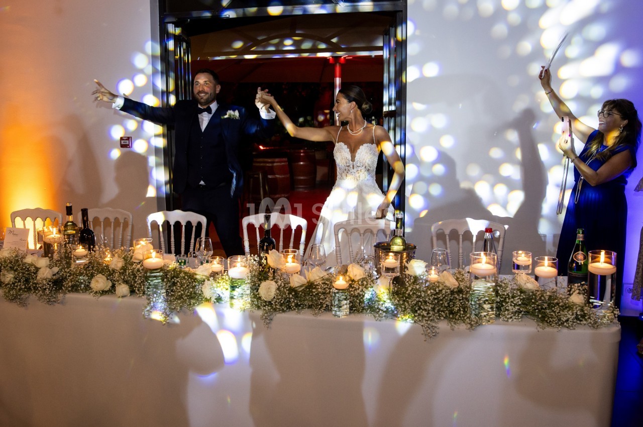 Un couple en tenue de mariage entre dans une salle décorée, accueilli par des invités et des jeux de lumière.