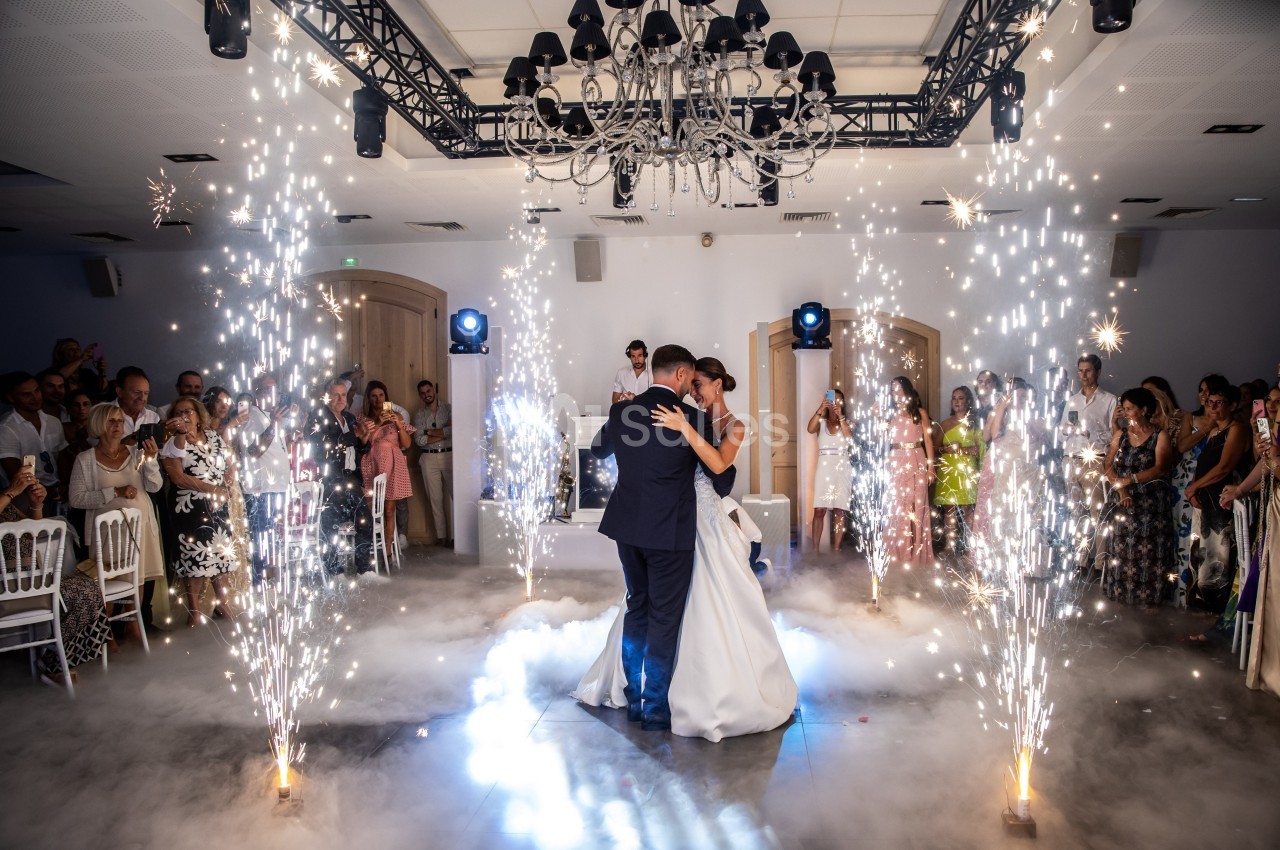 Un couple danse au centre d'une salle de réception, entouré de jets d'étincelles et de fumée légère.