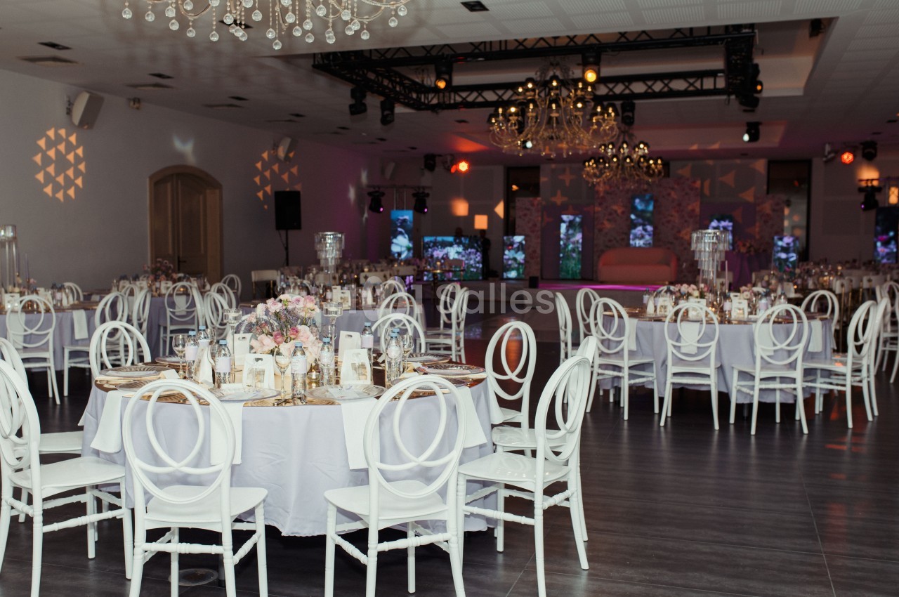 Salle de réception élégante avec tables rondes dressées, chaises blanches et décorations lumineuses suspendues.