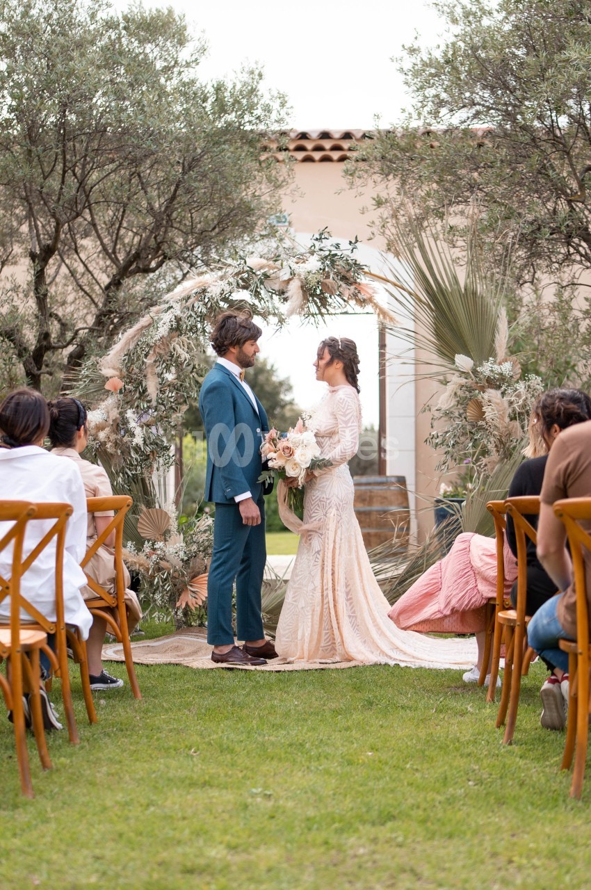 Un couple se tient devant une arche florale lors d'une cérémonie de mariage en extérieur, entouré d'invités assis.