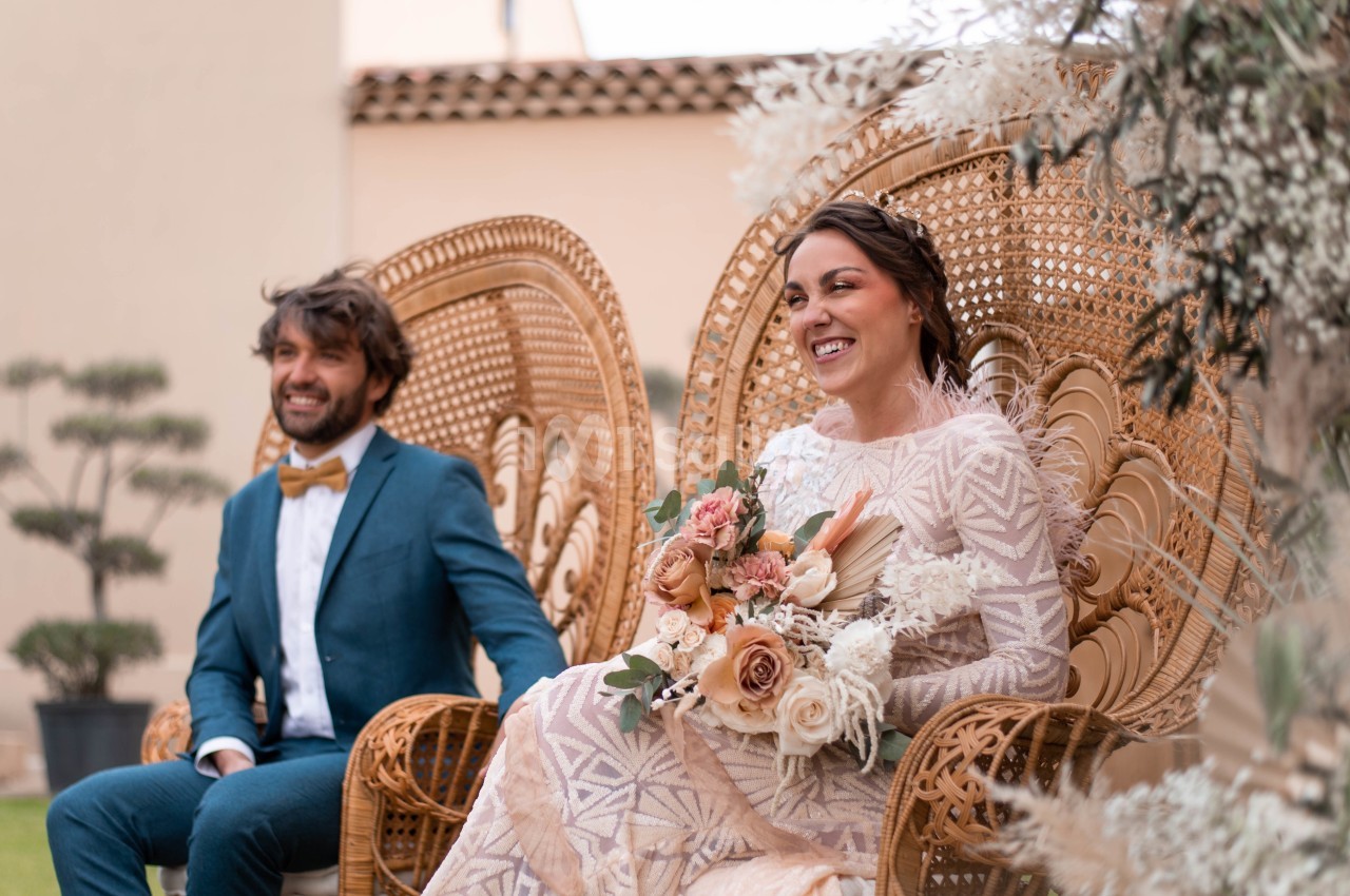 Un couple assis sur des fauteuils en rotin, la mariée tenant un bouquet de fleurs, dans un cadre extérieur lumineux.