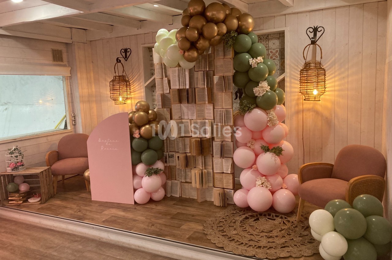 Décor intérieur avec arche de ballons pastel, livres suspendus, fauteuils roses et éclairage chaleureux.