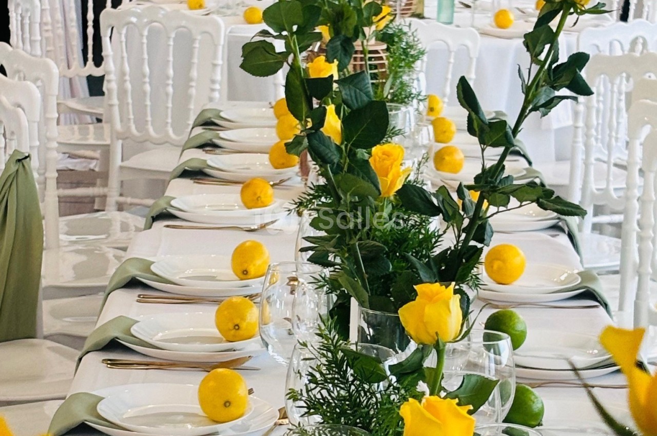 Table décorée avec des roses jaunes, des citrons et des éléments verts, entourée de chaises blanches.