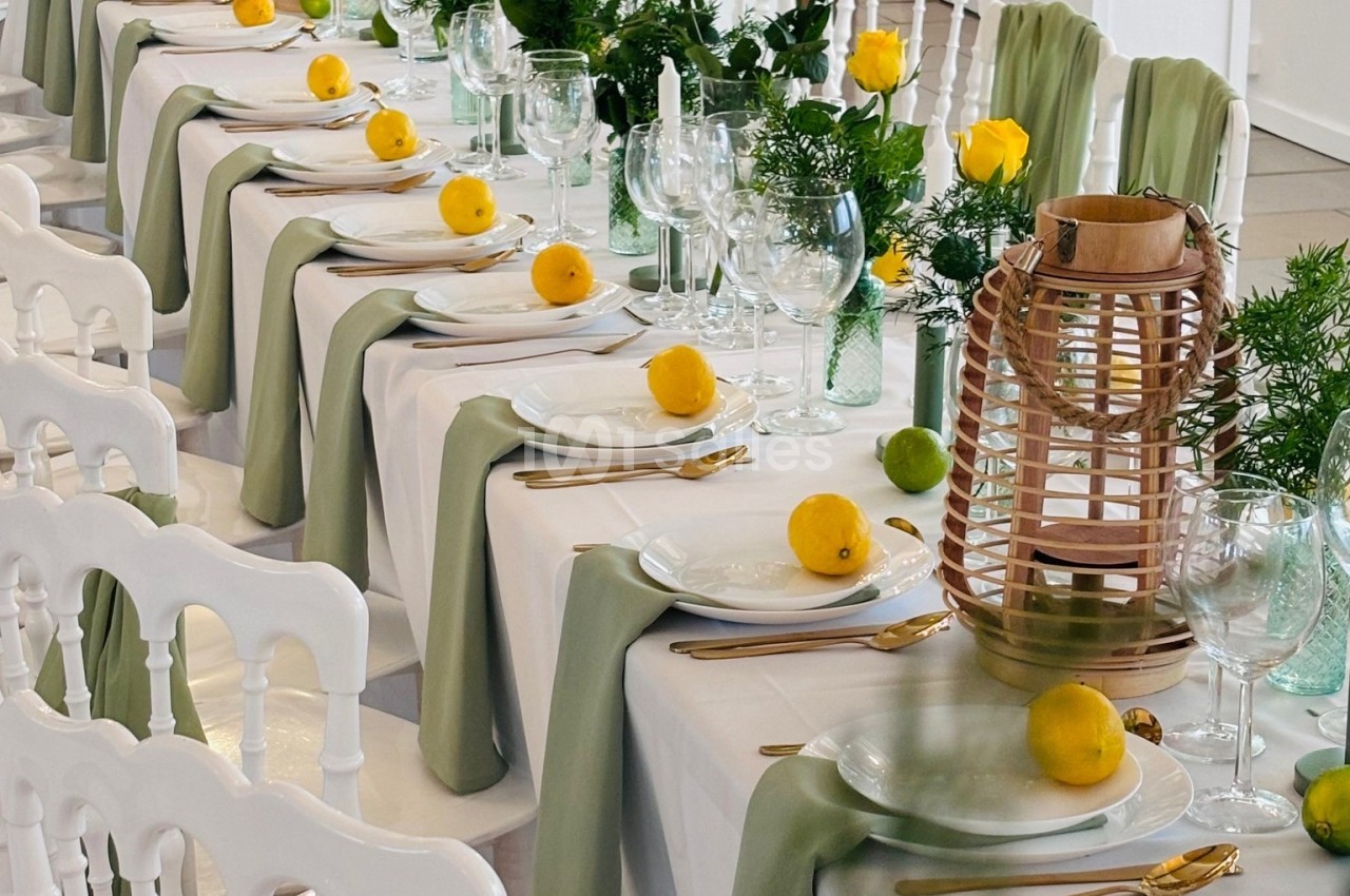 Table dressée avec nappes blanches, chemins de table verts, citrons jaunes, lanternes en osier et décorations florales.