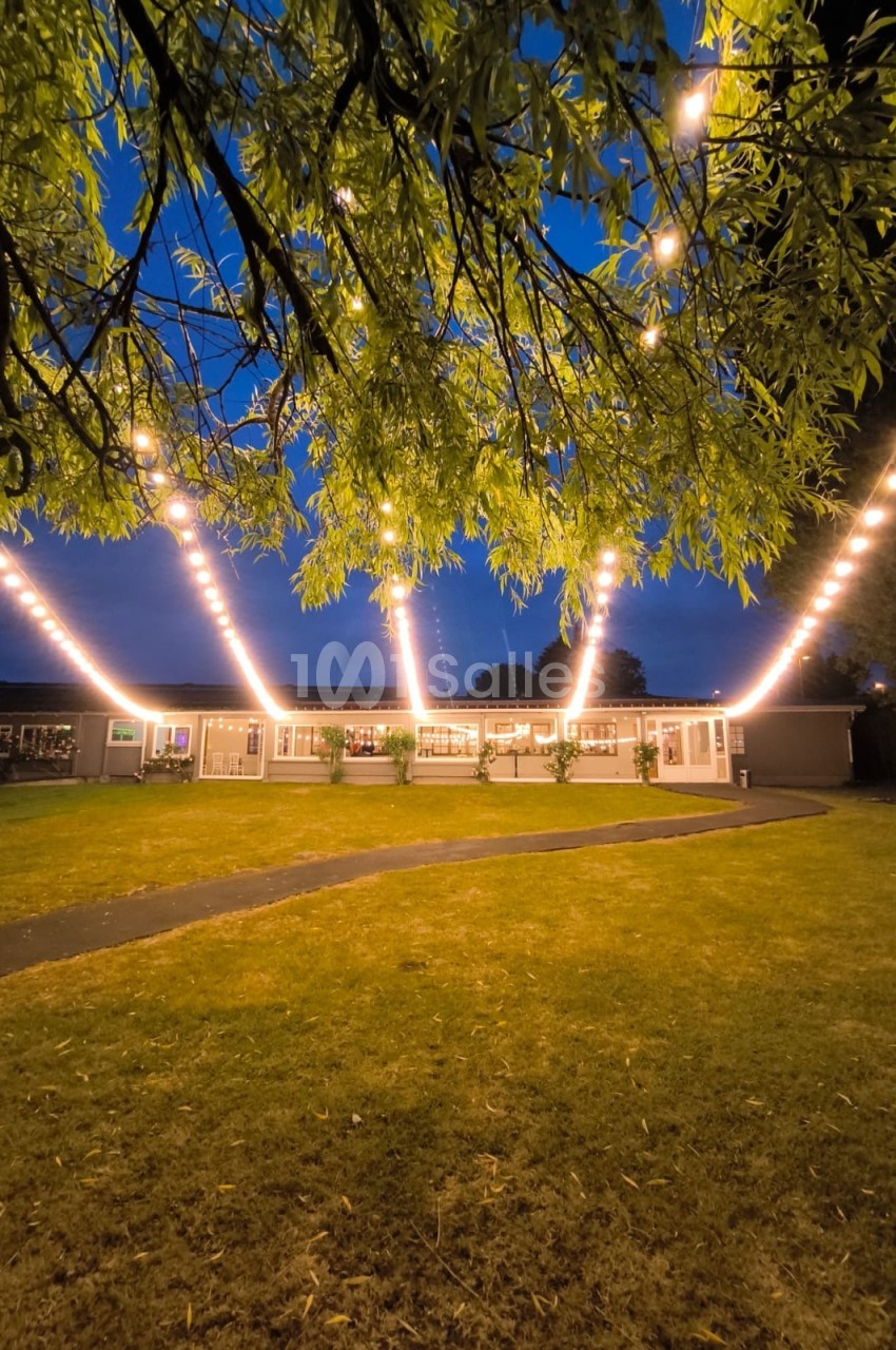 Guirlandes lumineuses suspendues au-dessus d'une pelouse devant un bâtiment éclairé, au crépuscule.