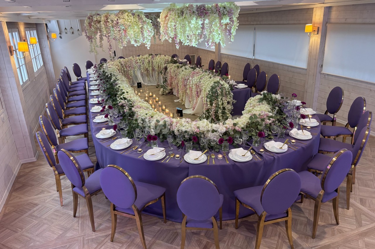 Salle de réception avec une grande table ovale décorée de fleurs blanches et violettes, nappes violettes et chaises…