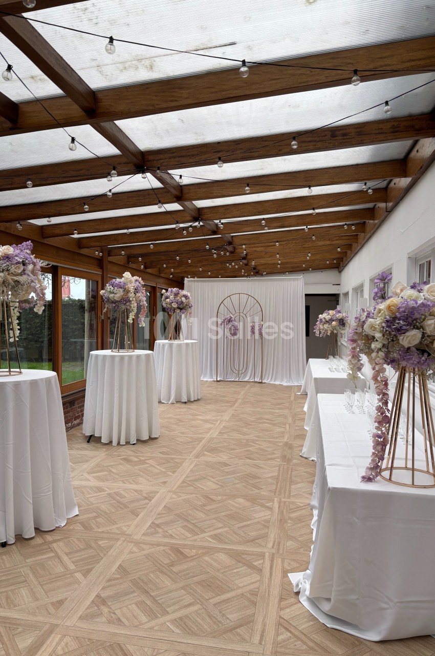 Salle décorée pour un événement, avec tables hautes drapées de blanc et ornées de bouquets pastel sous une verrière.