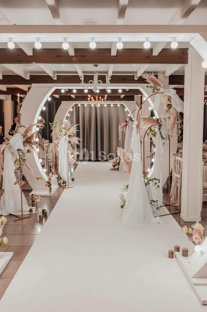 Allée décorée pour un mariage avec arches lumineuses, voilages blancs et arrangements floraux dans une salle élégante.