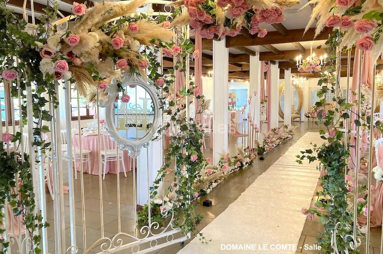 Allée décorée pour un mariage avec arche florale, roses, pampas et chemin blanc dans une salle élégante.
