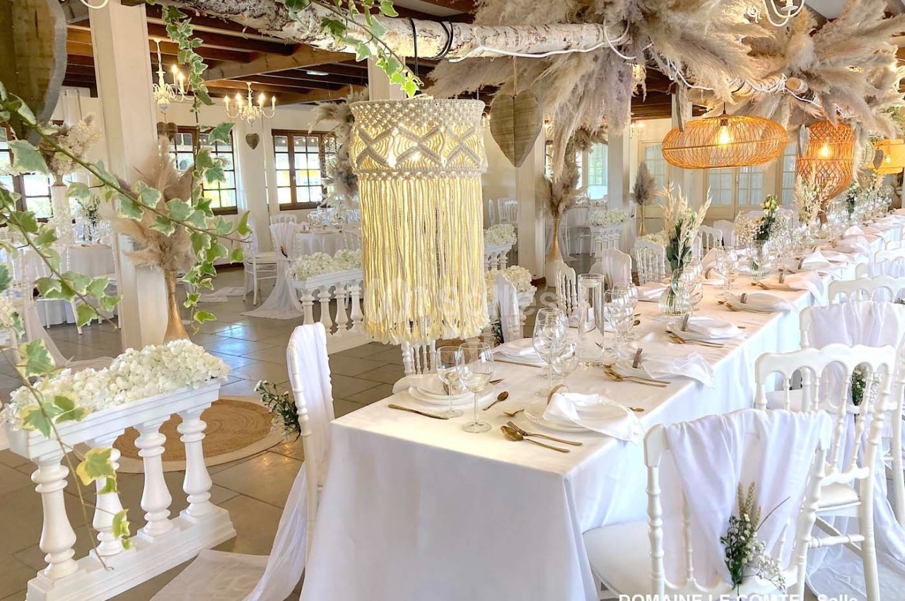 Salle de réception décorée avec des tables dressées, des chaises blanches et des ornements suspendus en fibres naturelles.