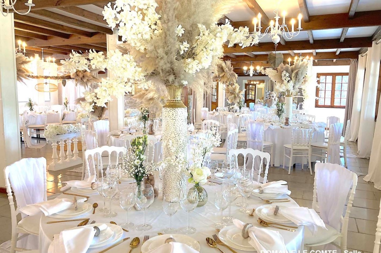Salle de réception décorée avec des tables rondes, nappes blanches, fleurs blanches et pampas, éclairée par des lustres.