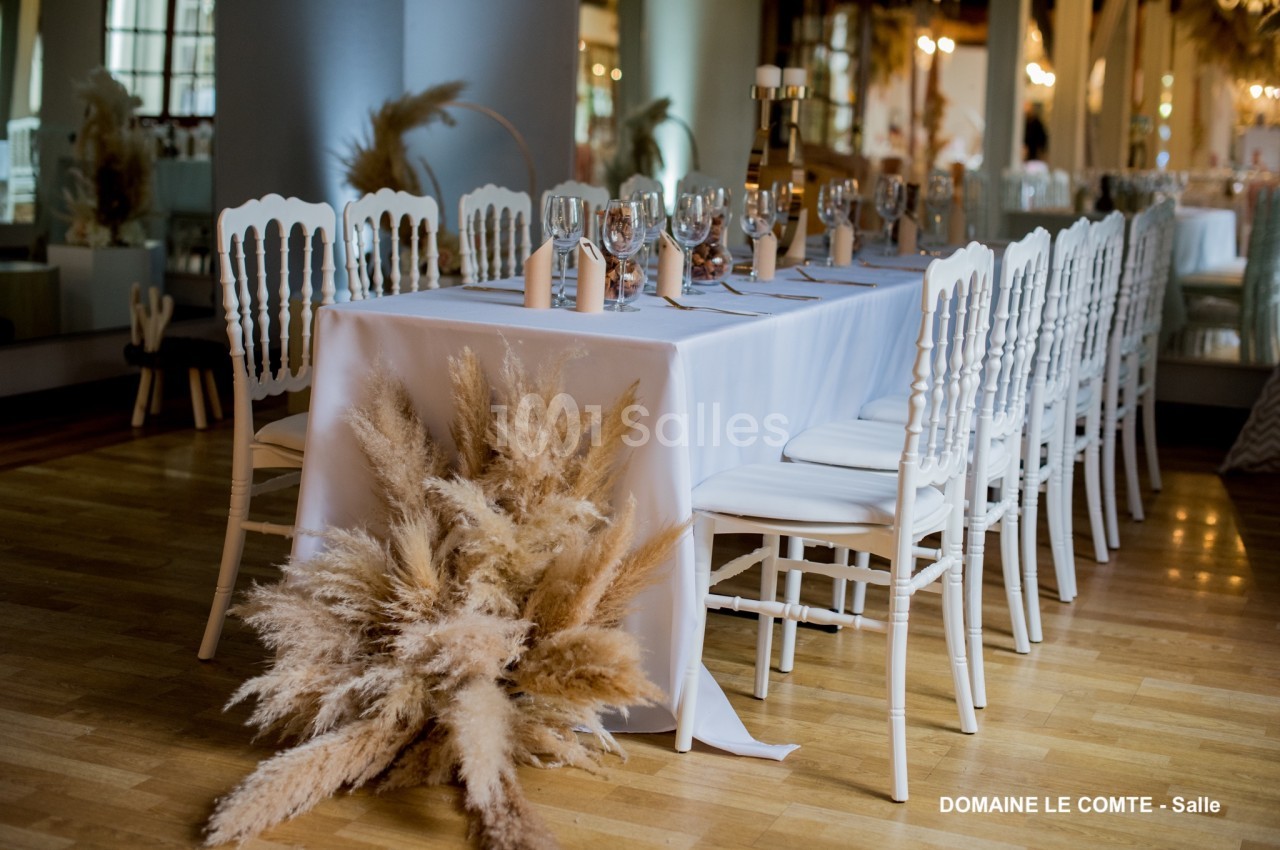 Table décorée avec nappes blanches, chaises assorties et bouquet de pampas, dans une salle lumineuse au sol en bois.
