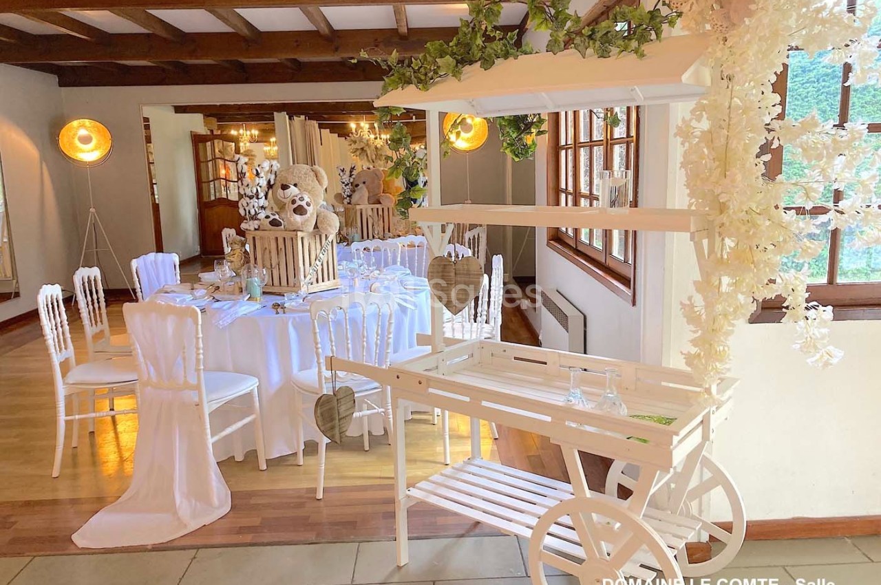 Salle de réception décorée avec des tables dressées, un chariot blanc orné de fleurs et une ambiance lumineuse chaleureuse.
