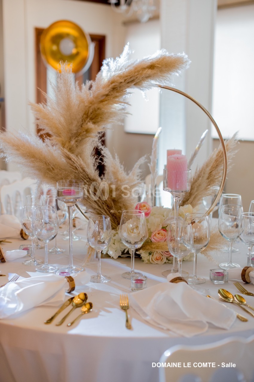 Table décorée pour un événement avec des pampas, des roses, un miroir rond, des bougies roses et des couverts dorés.