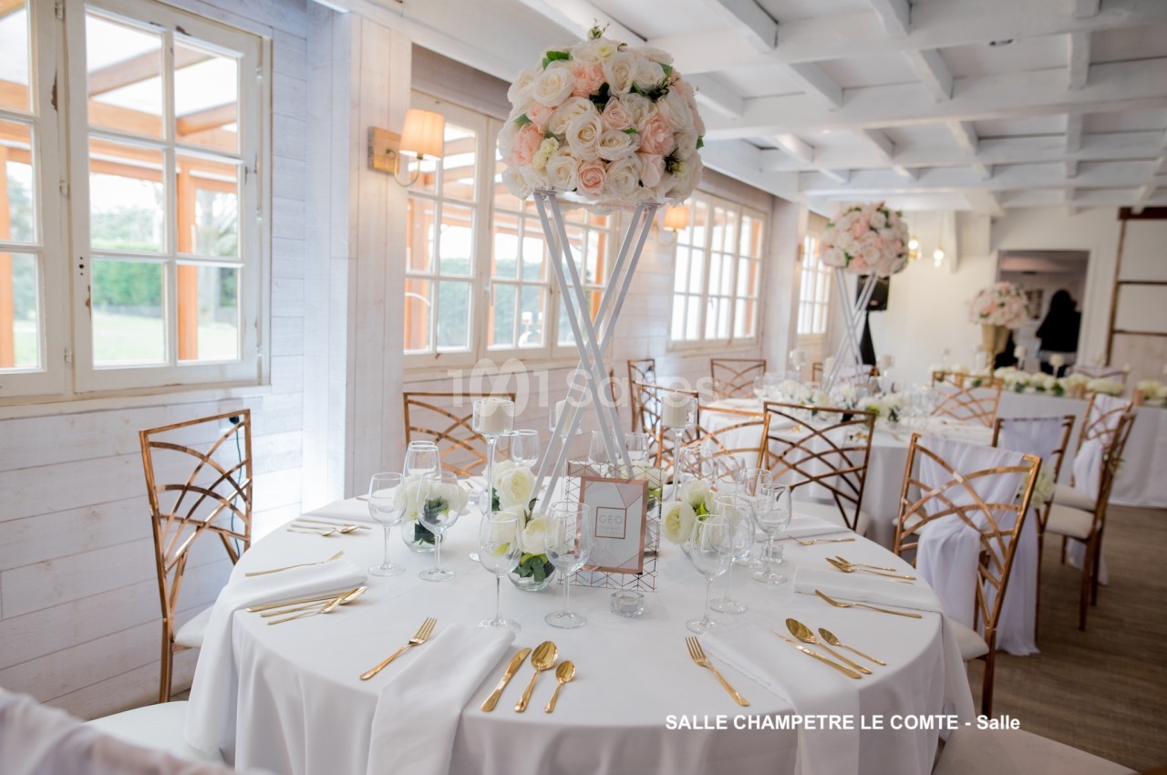 Salle de réception lumineuse avec tables rondes décorées de nappes blanches, fleurs et couverts dorés.