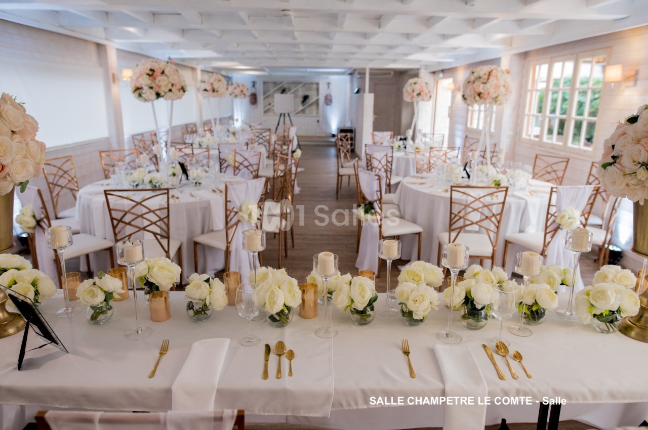 Salle de réception décorée avec des tables rondes, nappes blanches, chaises dorées et arrangements floraux élégants.