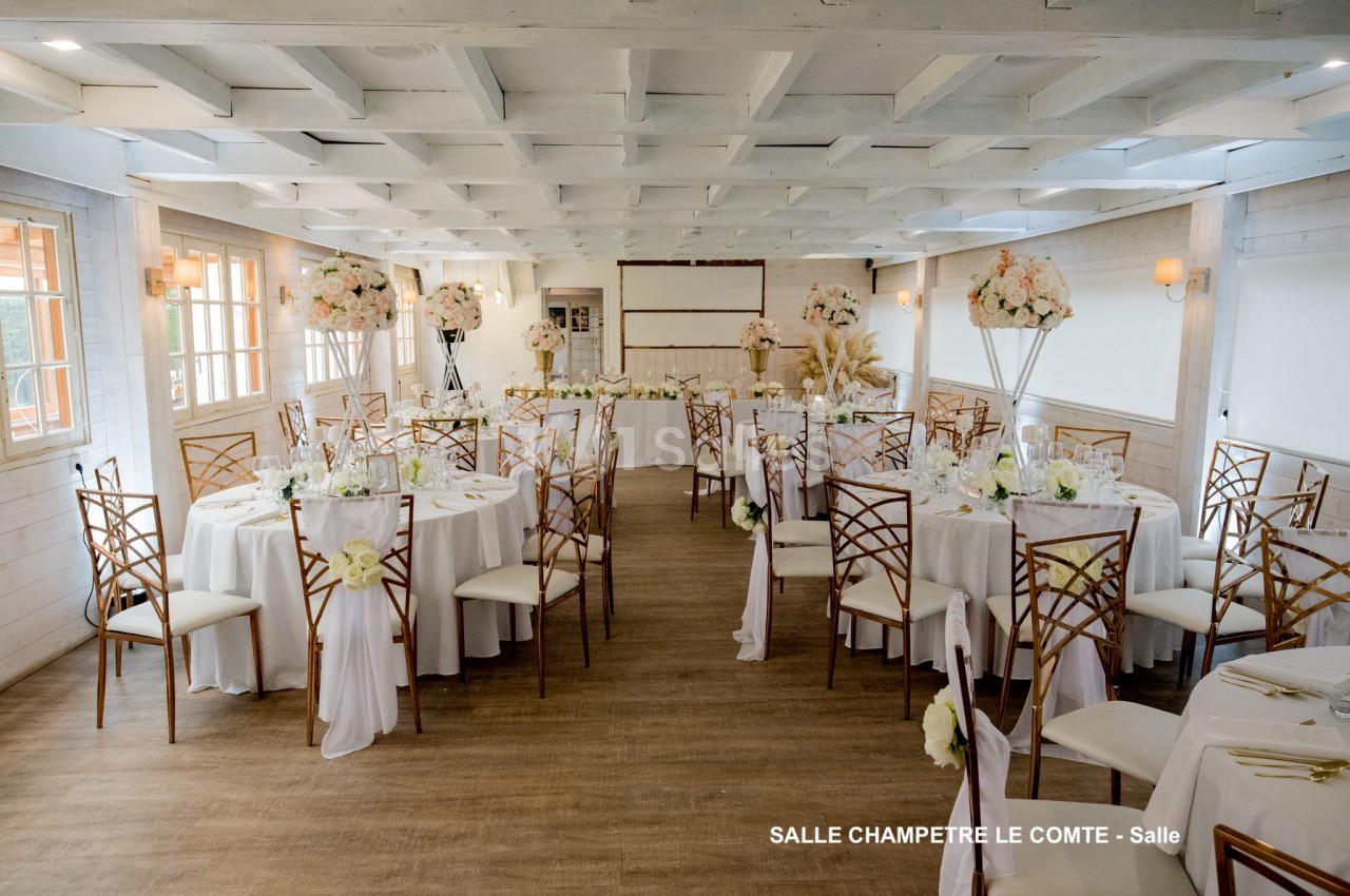 Salle de réception lumineuse avec tables rondes décorées de nappes blanches, chaises en bois et arrangements floraux.