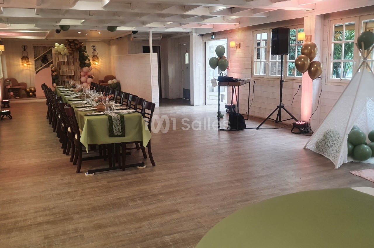 Salle décorée pour un événement, avec une longue table dressée, ballons colorés et un espace détente avec tipi.