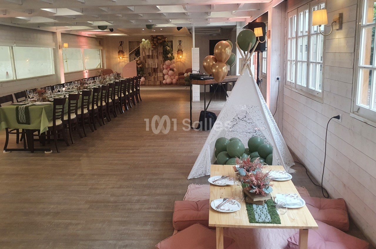 Salle décorée pour un événement avec une grande table dressée, un tipi, des ballons et une table basse avec coussins.