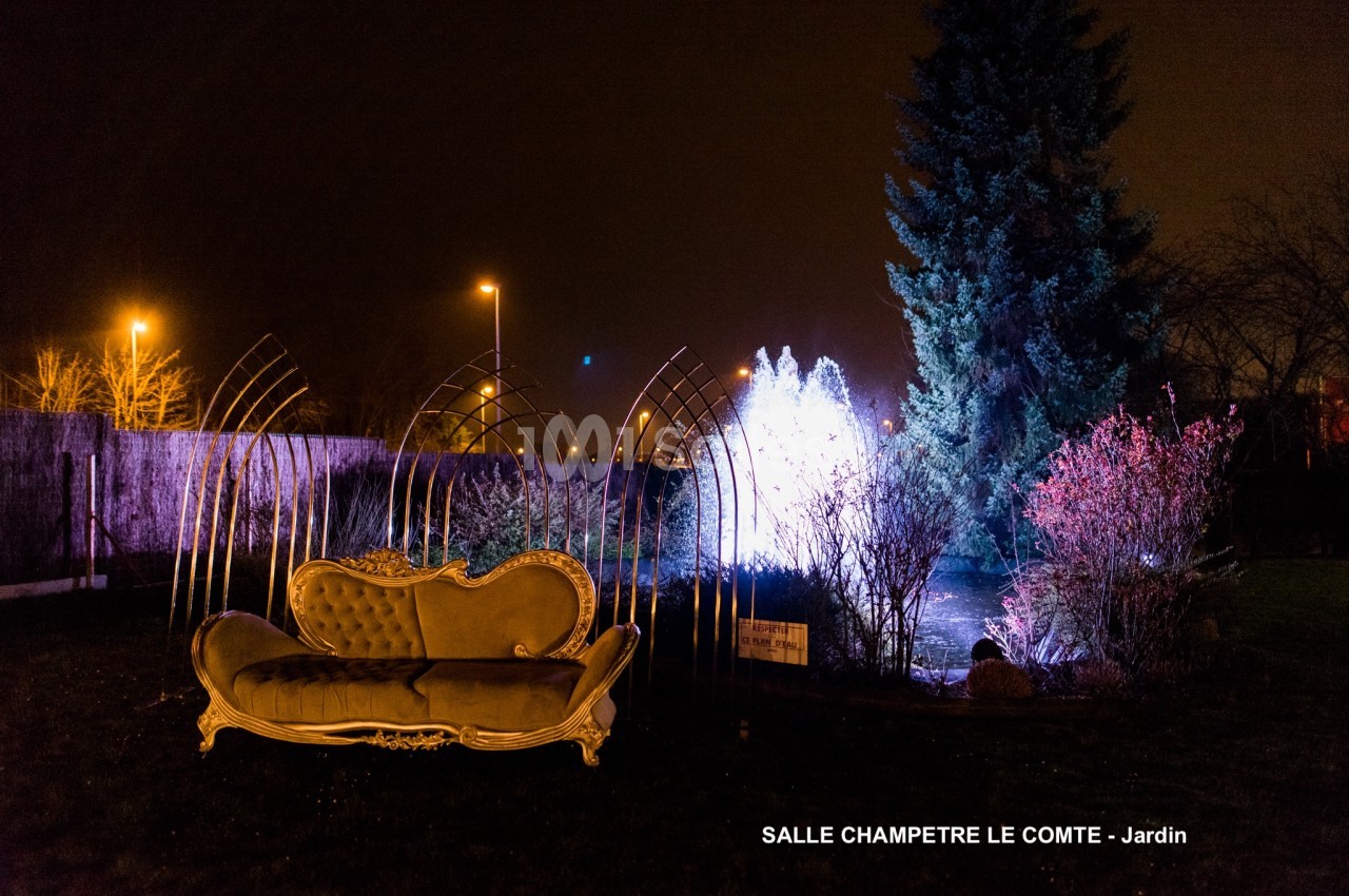 Canapé ancien doré dans un jardin nocturne éclairé par des lumières colorées, avec arbres et décorations métalliques.