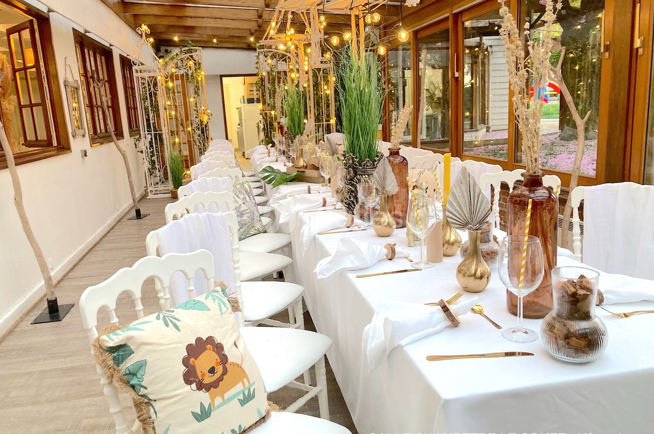 Salle décorée pour un événement, avec tables alignées, nappes blanches, chaises blanches et décorations végétales.