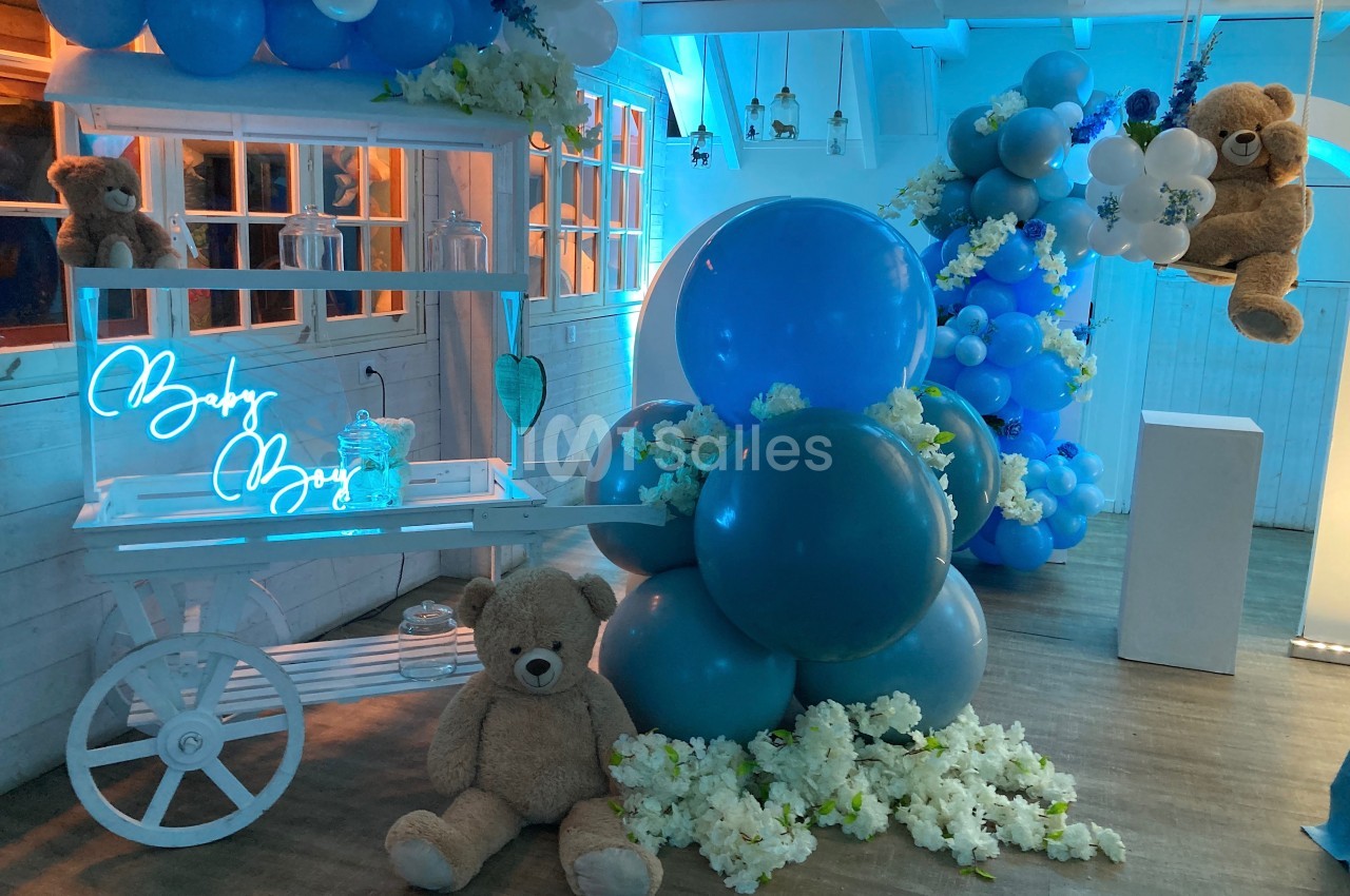 Décoration de fête avec ballons bleus, ours en peluche et fleurs blanches dans une ambiance lumineuse bleutée.
