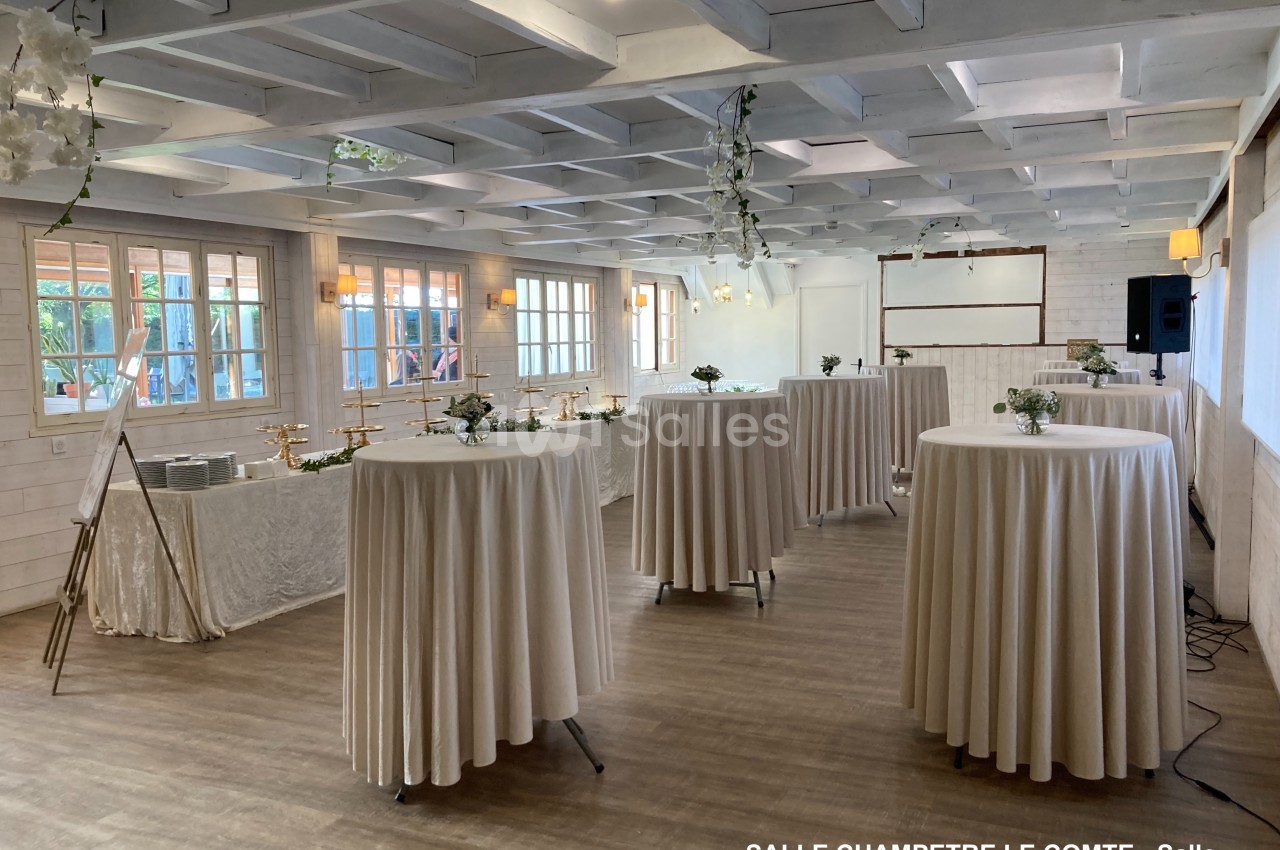 Salle lumineuse avec tables hautes drapées de blanc, décorée de fleurs, parquet au sol et poutres apparentes au plafond.