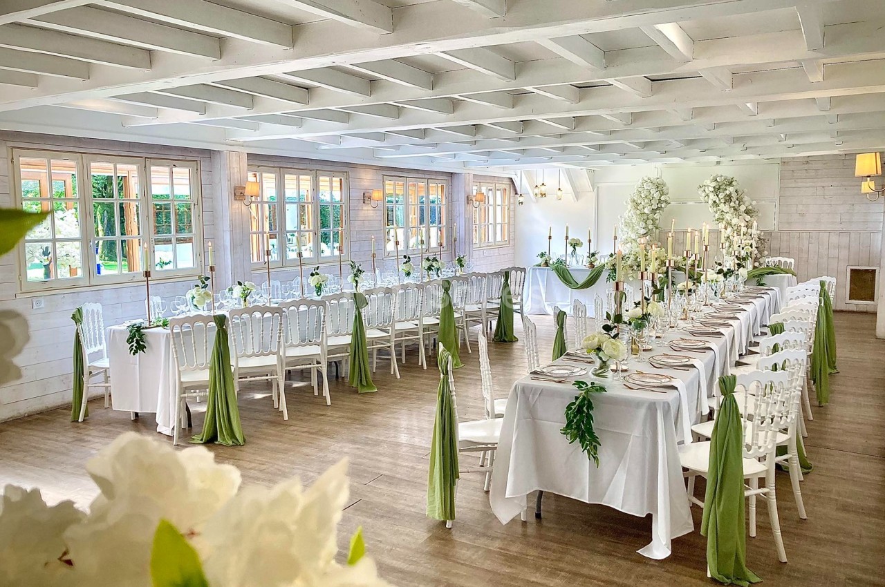 Salle de réception lumineuse décorée pour un mariage, avec longues tables ornées de nappes blanches et touches de vert.