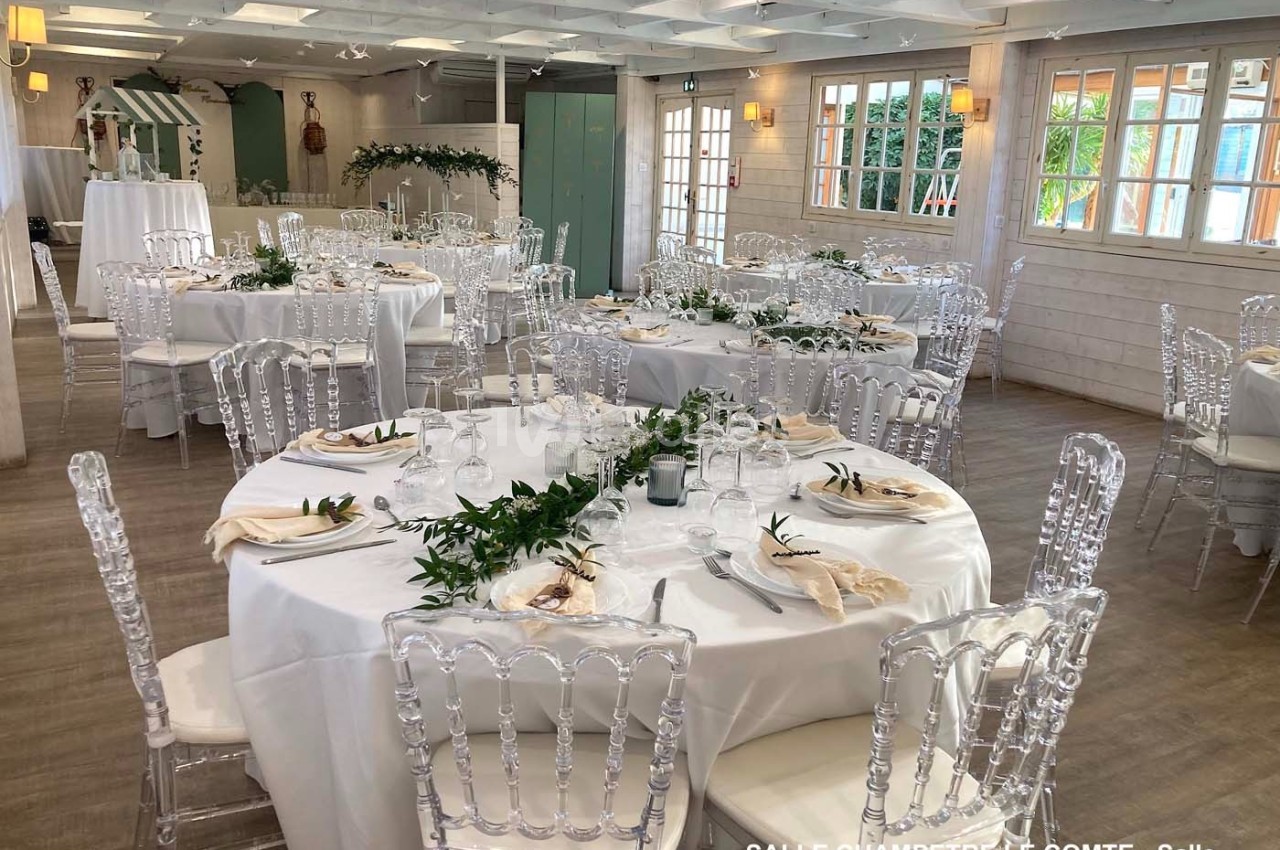 Salle décorée pour un événement, avec tables rondes dressées, nappes blanches, chaises transparentes et ornements végétaux.