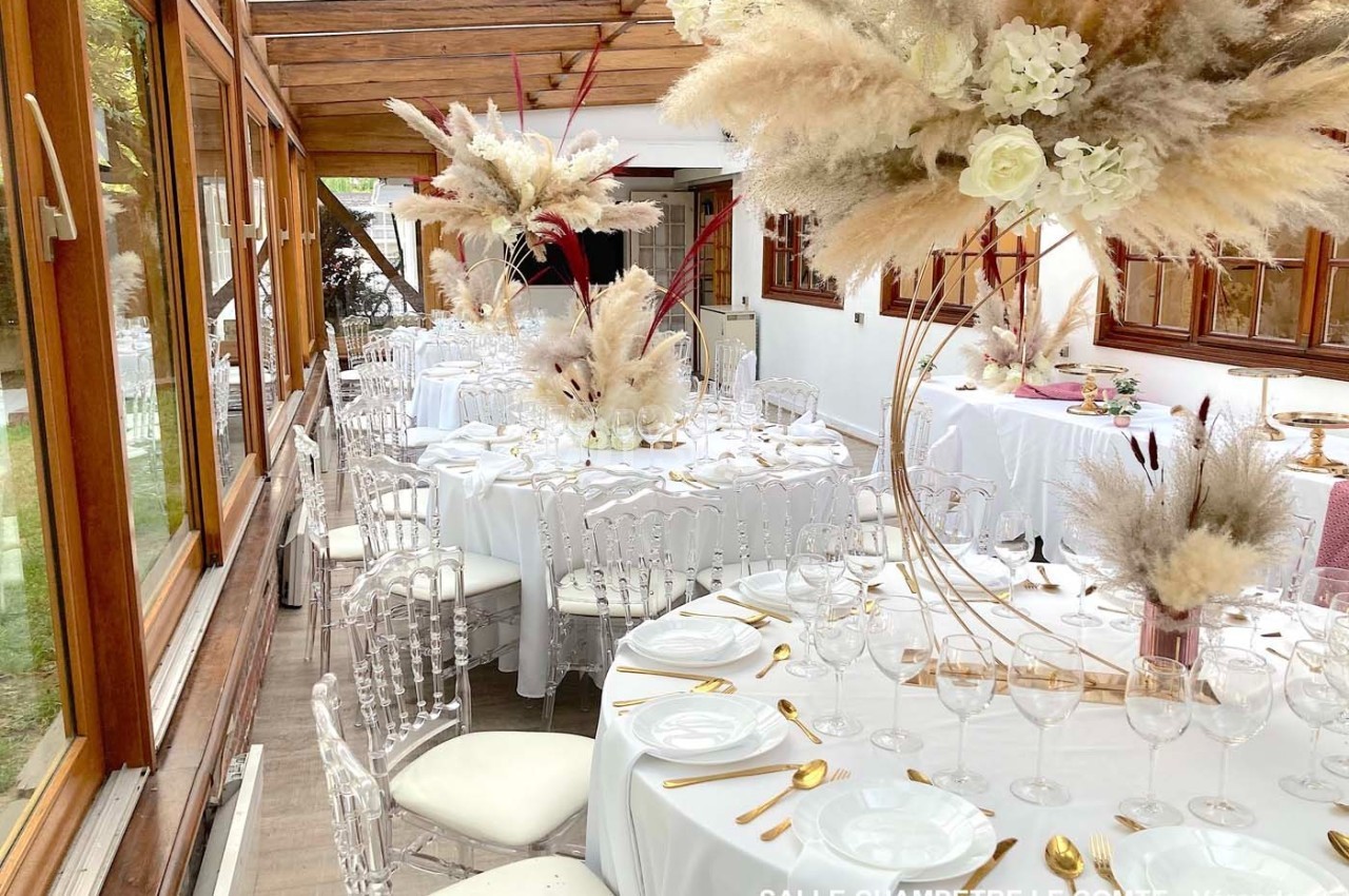 Salle décorée pour un événement, avec tables rondes dressées, chaises transparentes et arrangements floraux élégants.