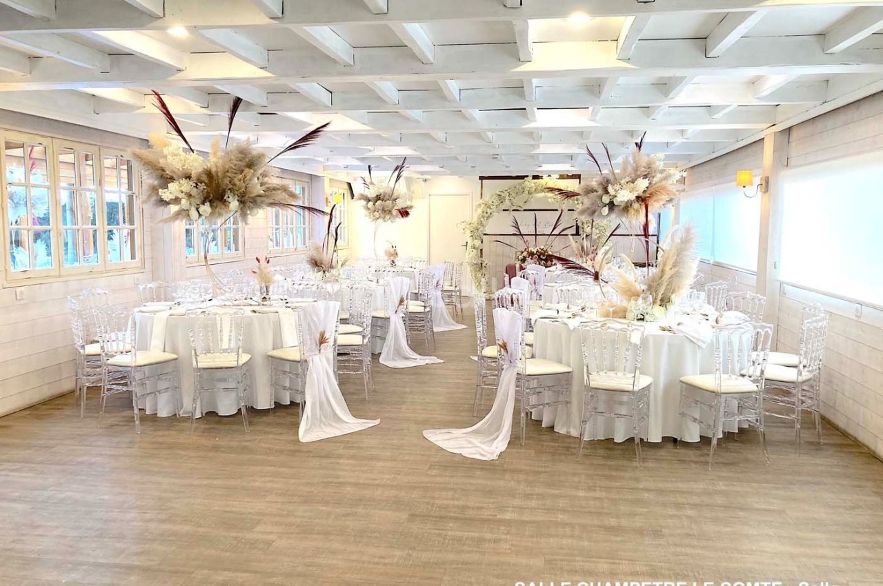 Salle de réception décorée avec des tables rondes, nappes blanches, chaises transparentes et arrangements floraux élégants.