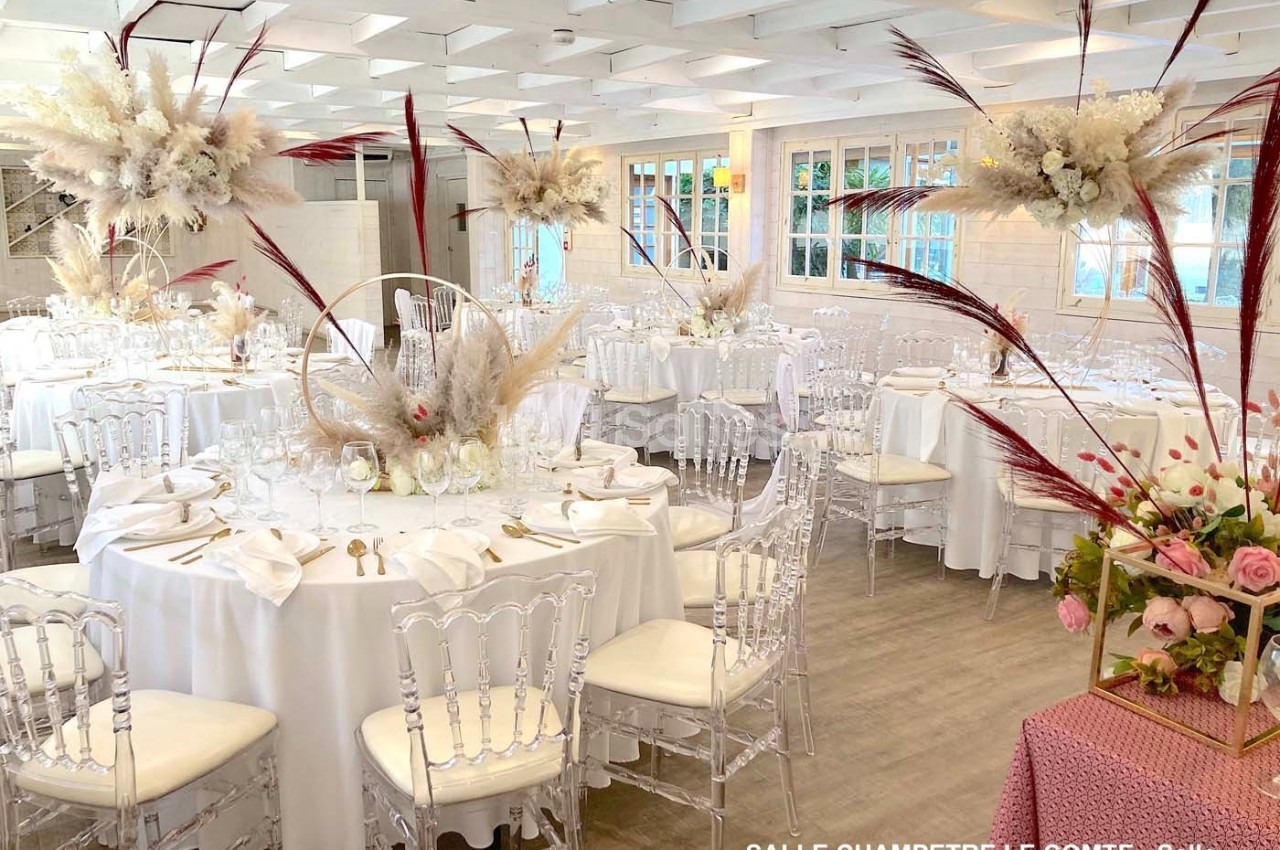 Salle de réception lumineuse décorée de tables rondes avec nappes blanches, chaises transparentes et compositions florales…