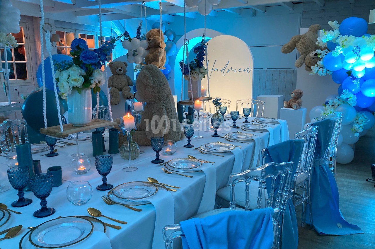 Table décorée pour une fête avec vaisselle élégante, ours en peluche, ballons bleus et bougies dans une ambiance lumineuse.