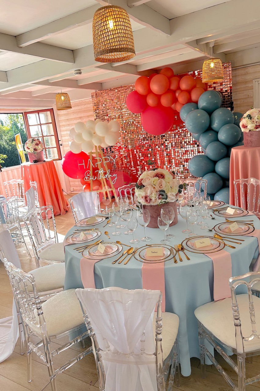 Salle décorée pour une fête avec des tables dressées, des ballons colorés et un mur scintillant en arrière-plan.