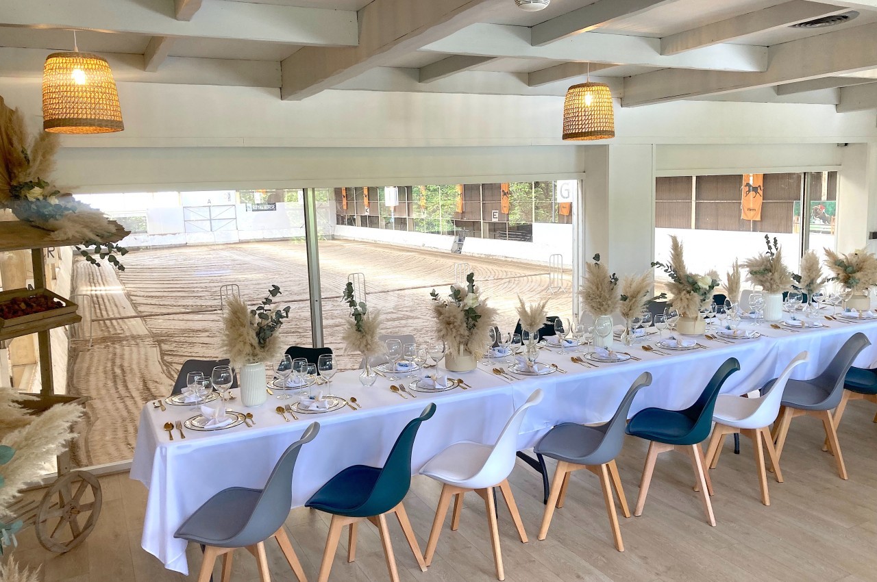 Salle lumineuse avec une longue table dressée, décorée de fleurs séchées, donnant sur une carrière équestre.