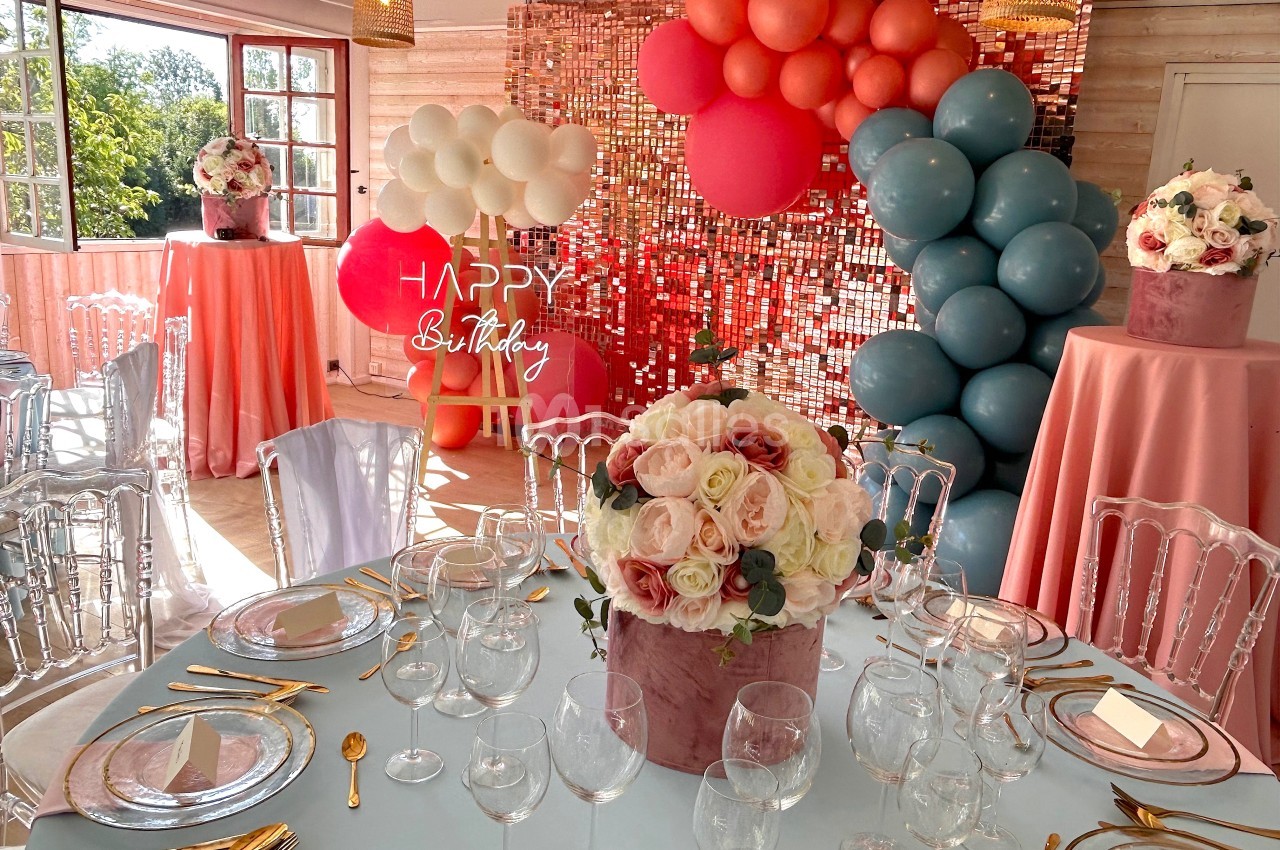 Salle décorée pour une fête avec ballons colorés, tables dressées et bouquets de fleurs au centre.