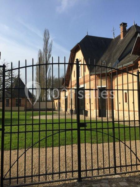 Vue d'une maison en briques avec toit en ardoise, entourée d'une pelouse et clôturée par une grille en métal noir.