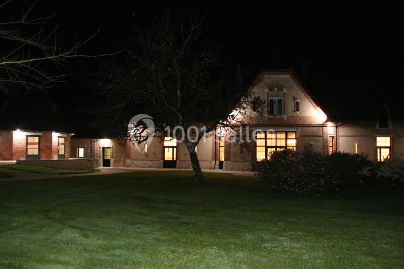 Bâtiment éclairé de nuit avec façade en pierre, fenêtres lumineuses et pelouse au premier plan.