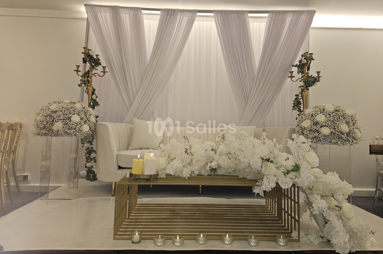 Décoration intérieure élégante avec canapé blanc, rideaux blancs, fleurs blanches et bougies sur une table dorée.