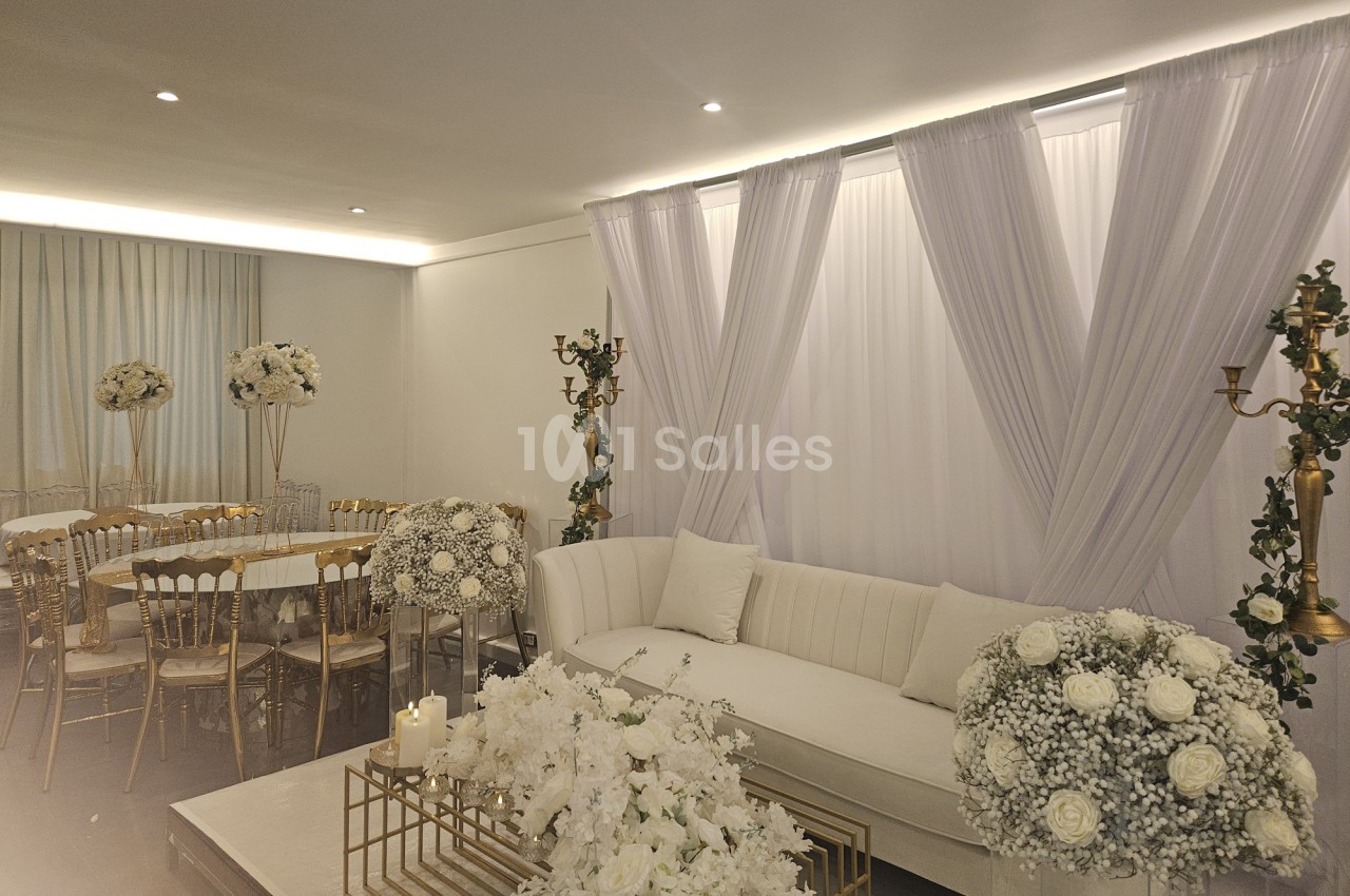 Salle décorée pour un événement avec canapés blancs, rideaux, chandeliers dorés et compositions florales blanches.