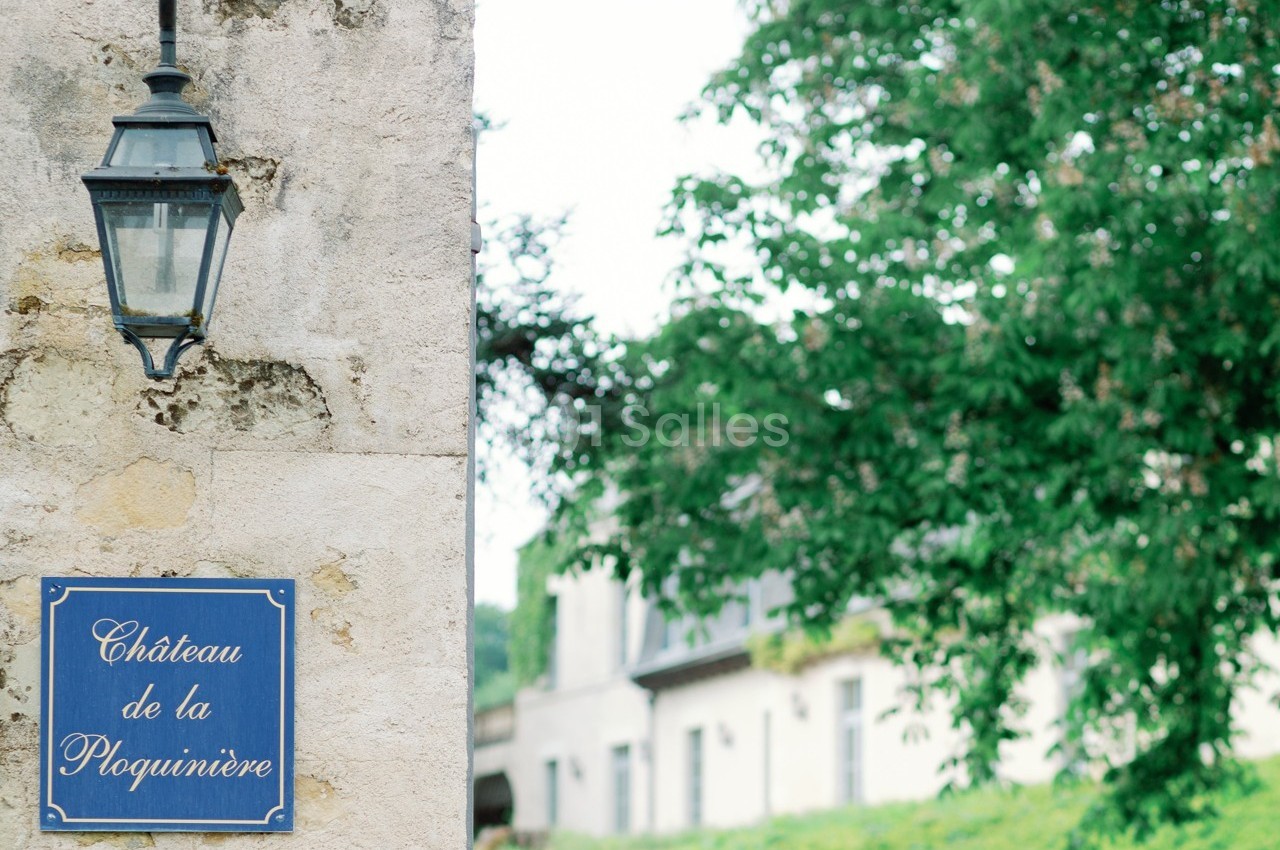 Plaque bleue indiquant ’Château de la Ploquinière’ fixée sur un mur en pierre, avec une lanterne et des arbres en arrière…