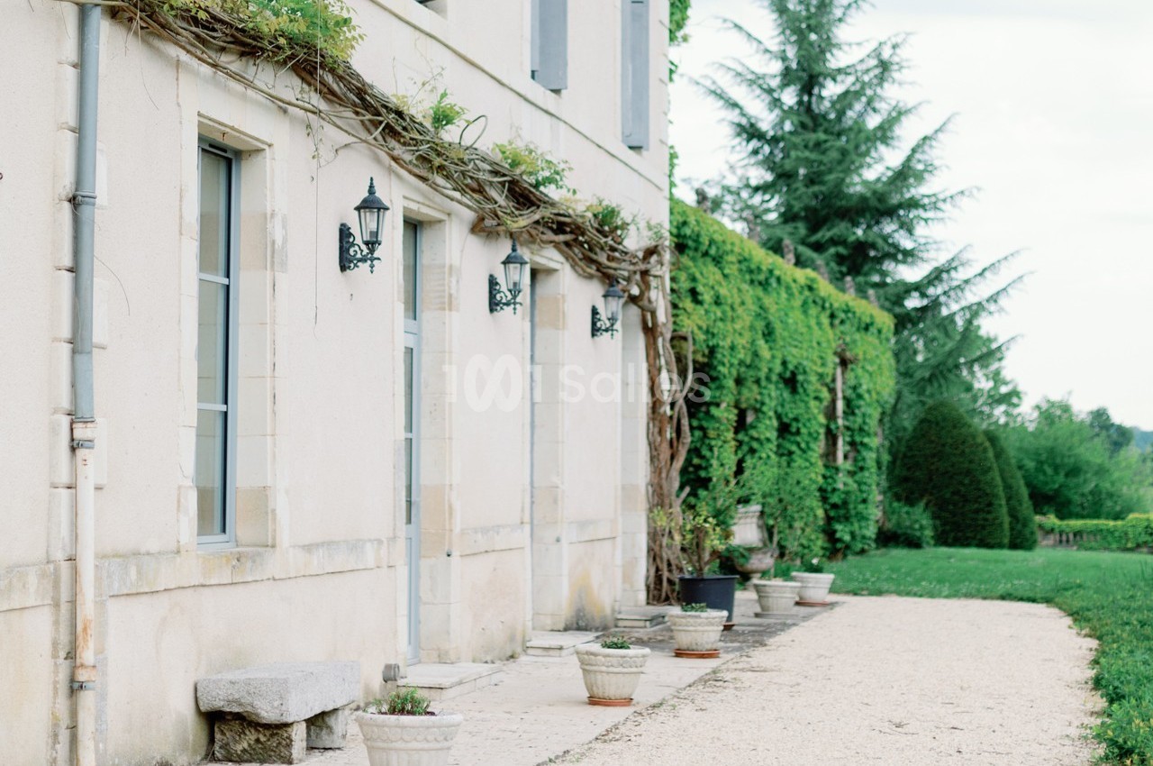 Façade d'une maison en pierre avec des lanternes murales, des plantes grimpantes et un chemin bordé de pots de fleurs.