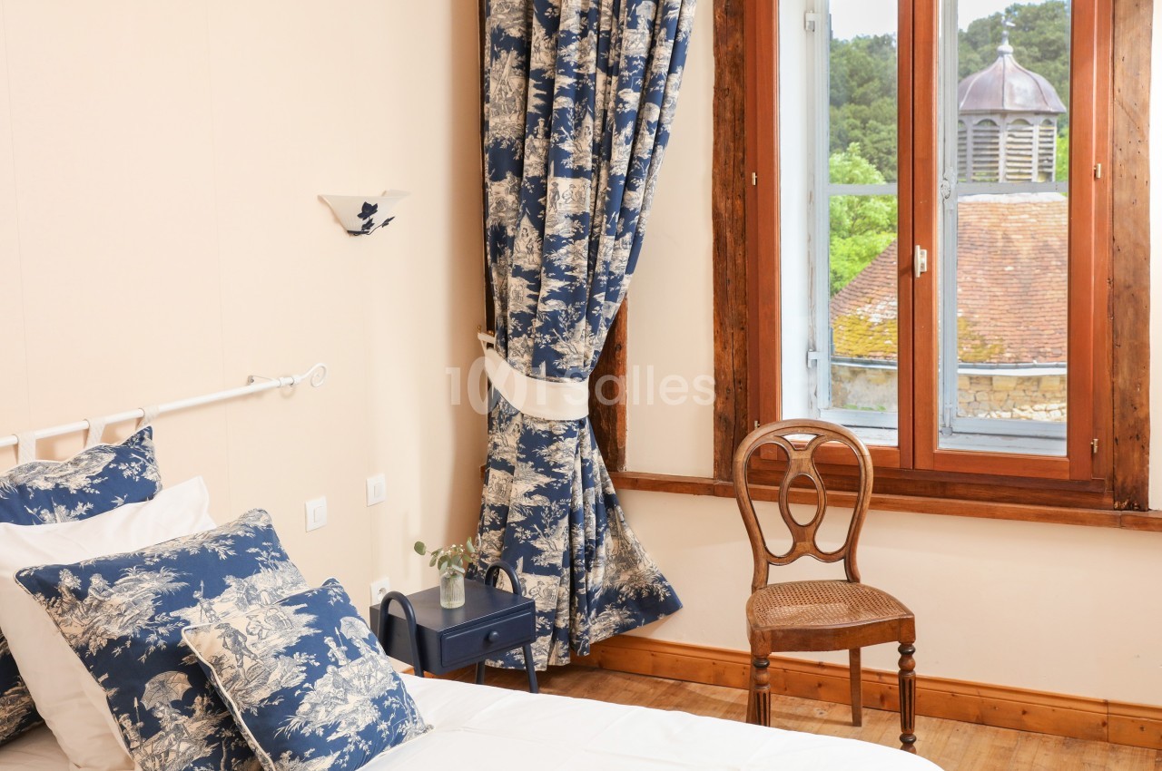 Chambre lumineuse avec lit, coussins et rideaux bleus à motifs, chaise en bois et fenêtre donnant sur un toit et des arbres.
