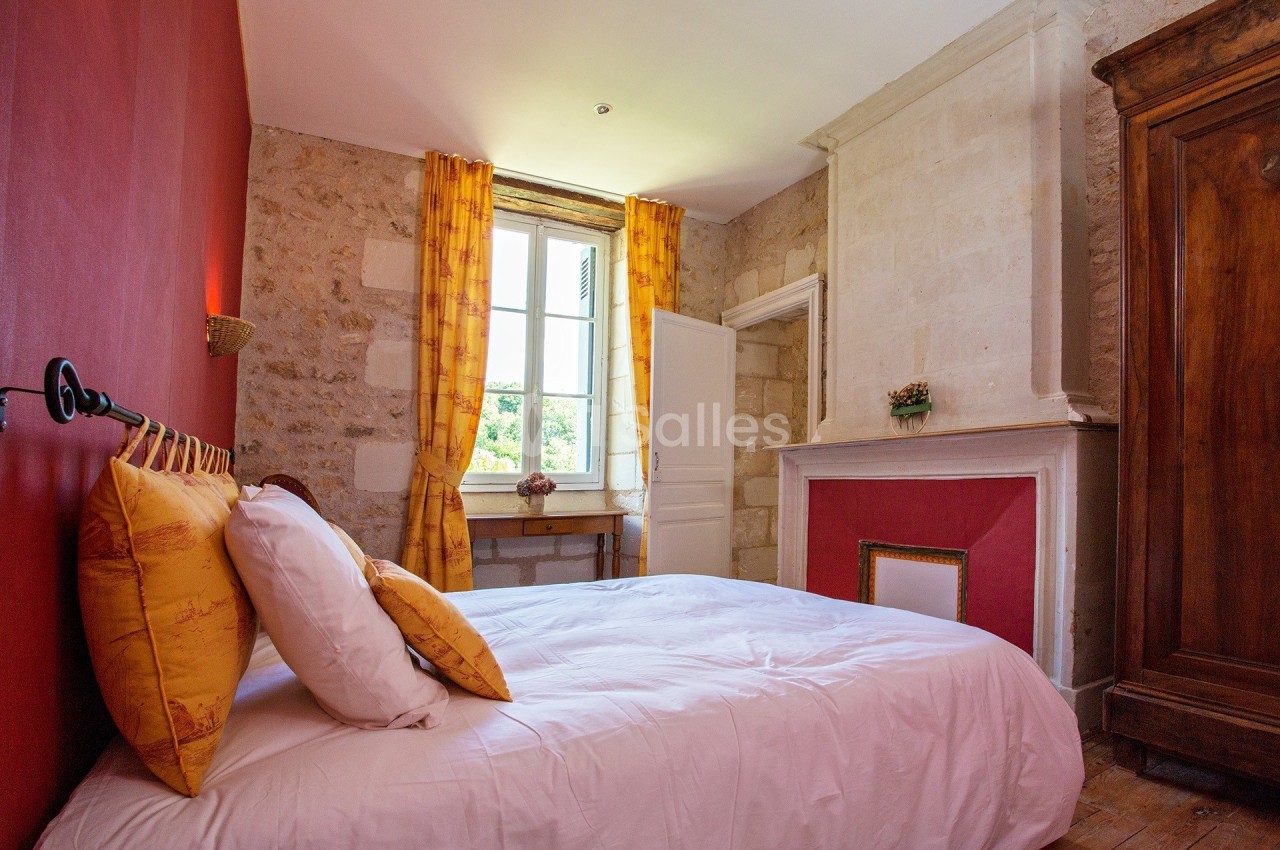Chambre lumineuse avec murs en pierre, cheminée blanche, rideaux jaunes et lit avec coussins assortis.