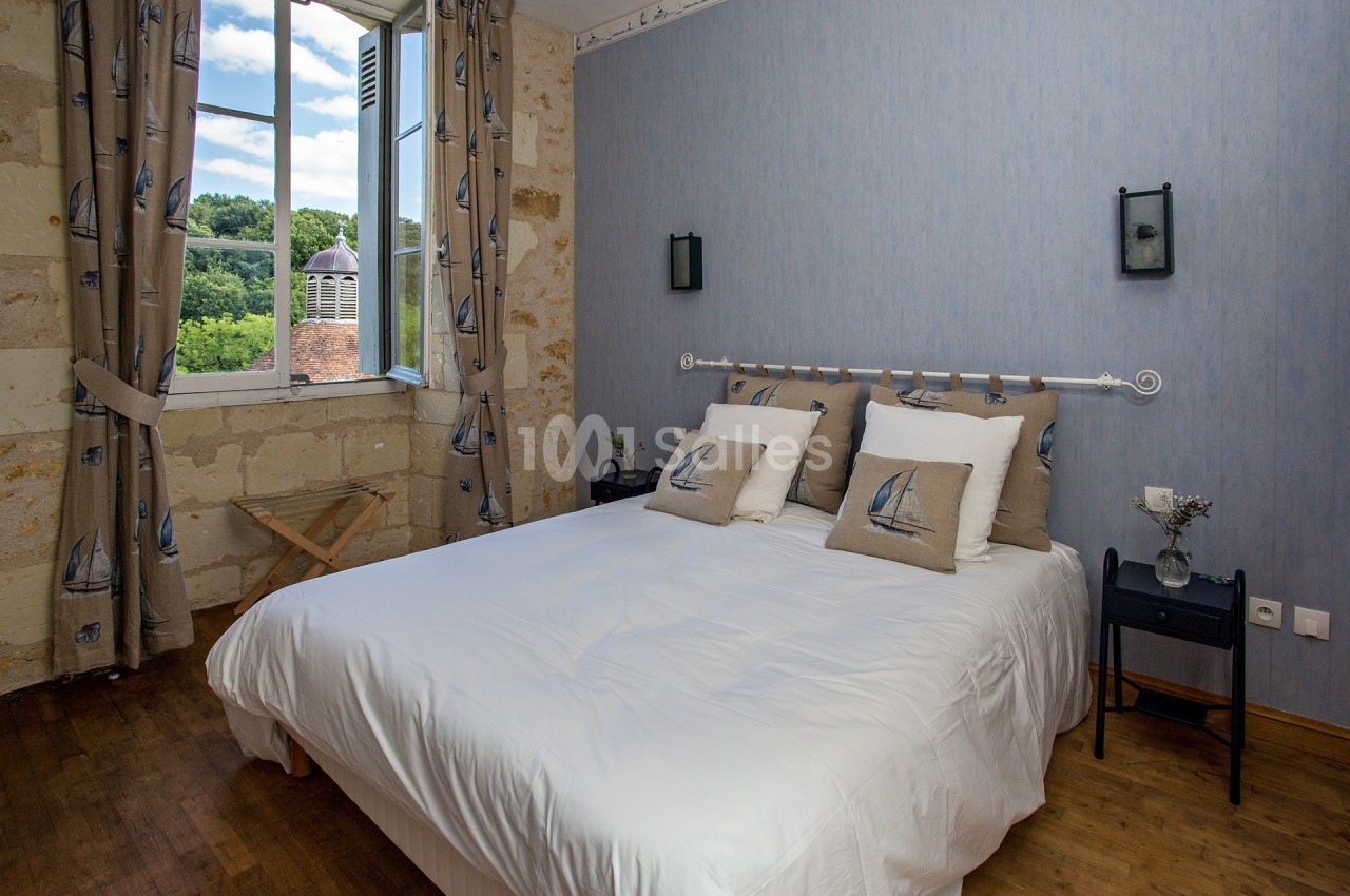 Chambre lumineuse avec un lit double, des coussins décorés de voiliers, et une fenêtre ouverte sur un paysage verdoyant.