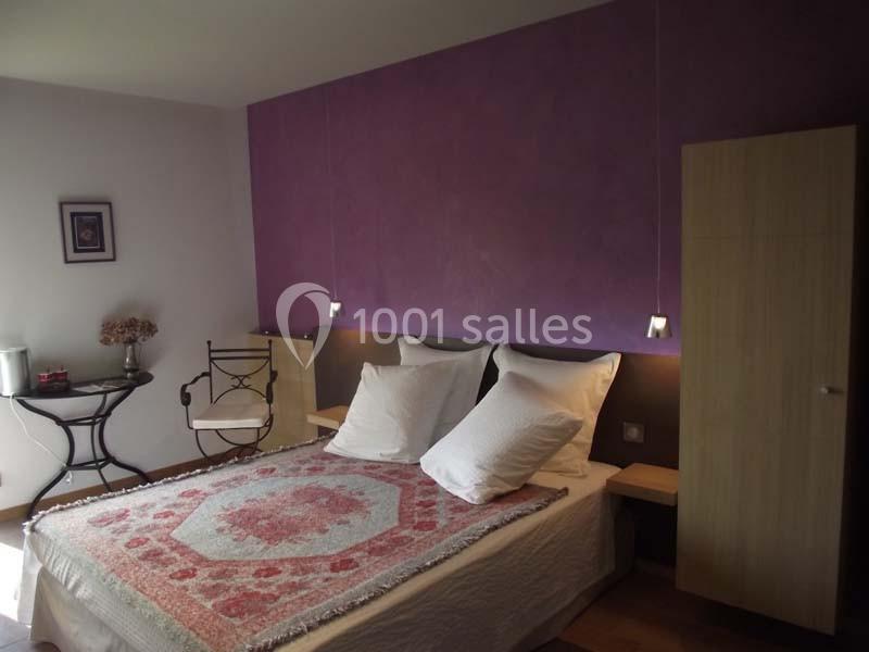 Chambre avec lit double, mur violet, table avec chaise en fer forgé, armoire en bois clair et décoration simple.