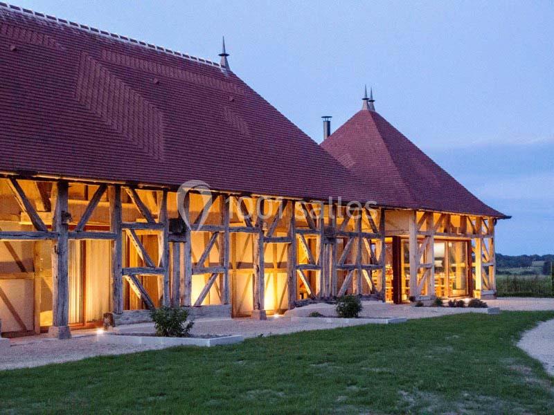 Bâtisse traditionnelle à colombages éclairée en soirée, entourée de pelouse et située dans un cadre rural.