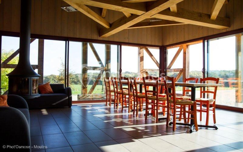 Salle lumineuse avec grandes baies vitrées, table en bois entourée de chaises et poêle à bois dans un coin.