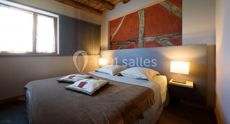 Chambre avec lit double, tête de lit en bois, éclairage doux et décoration murale avec poutres apparentes.