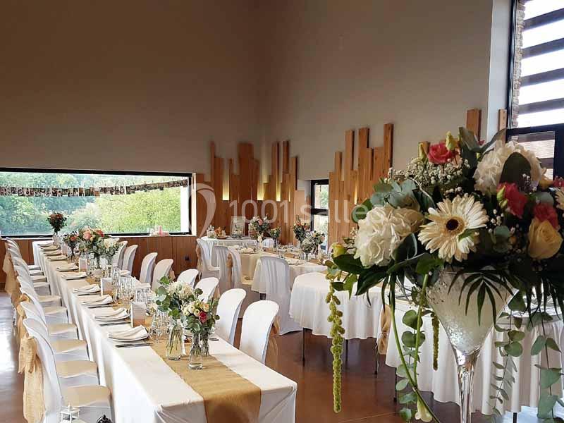 Salle de réception décorée avec des tables alignées, nappes blanches, fleurs et éléments en bois sur les murs.